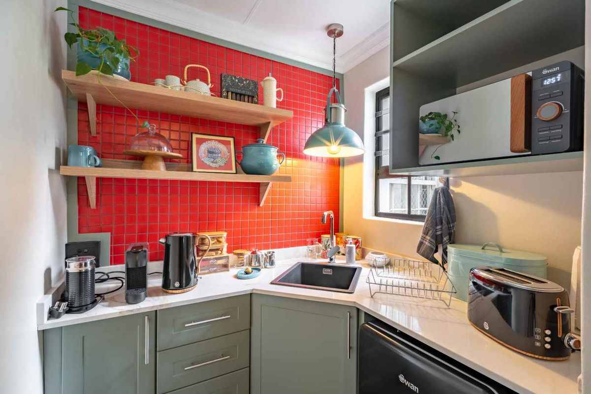 Compact kitchen featuring sage green cabinetry, stone countertops, a striking red tiled splashback, and floating timber shelving with integrated lighting.