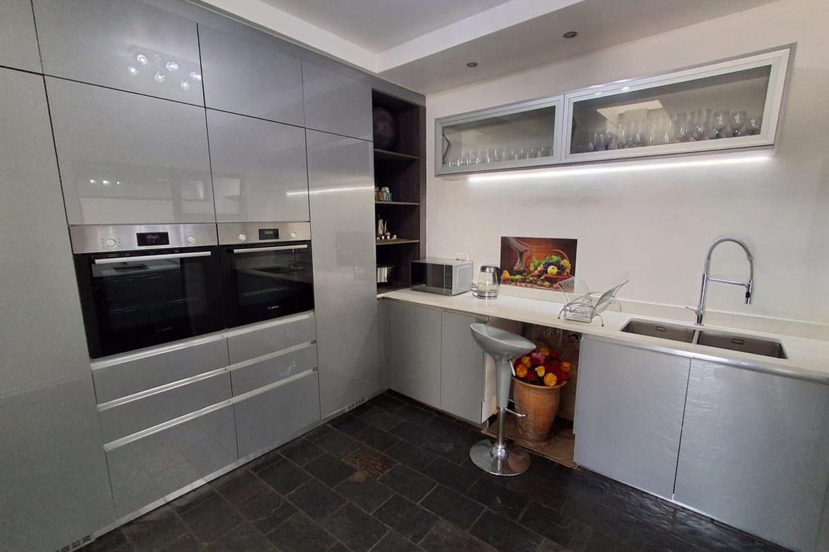 Modern kitchen featuring high-gloss grey cabinetry, dual built-in ovens, stone countertops, under-cabinet lighting, and dark slate flooring.