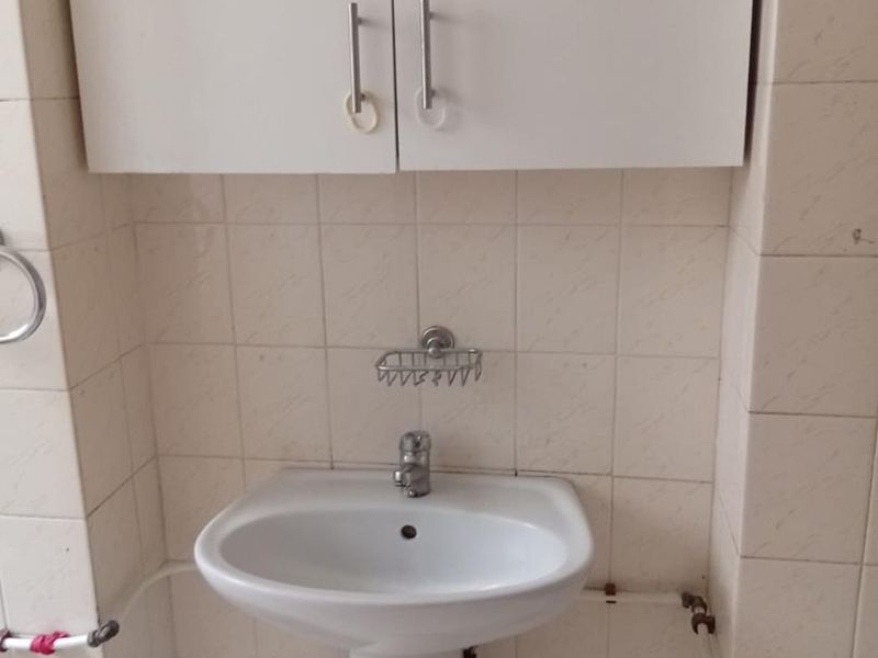 A compact bathroom featuring a white pedestal basin, wall-mounted storage cabinet, and tiled walls, requiring minor cosmetic updates to the exposed plumbing and flooring.