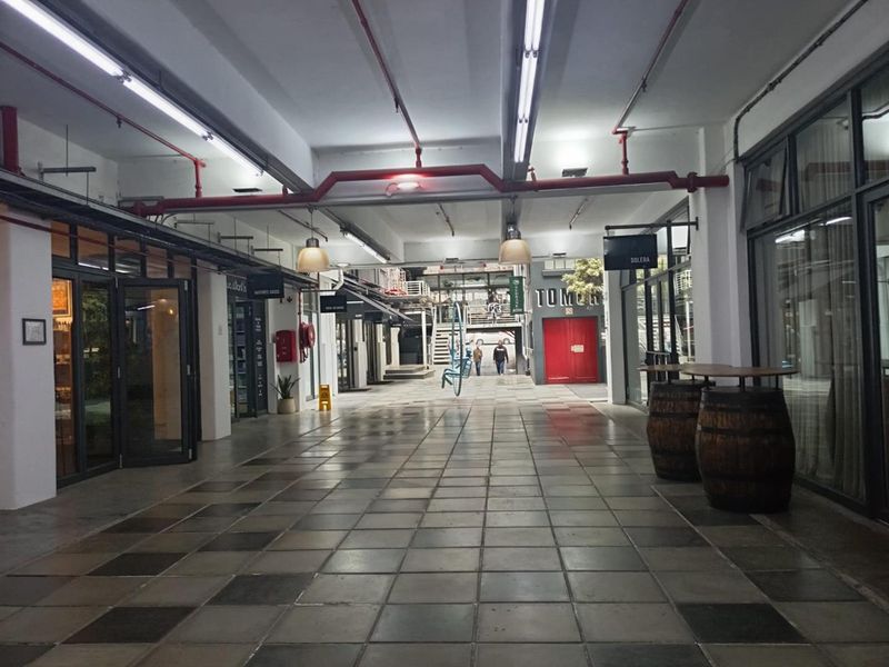 A commercial hallway or pedestrian thoroughfare featuring industrial-style exposed red fire piping, fluorescent lighting, and tiled flooring with access to various office or retail units.