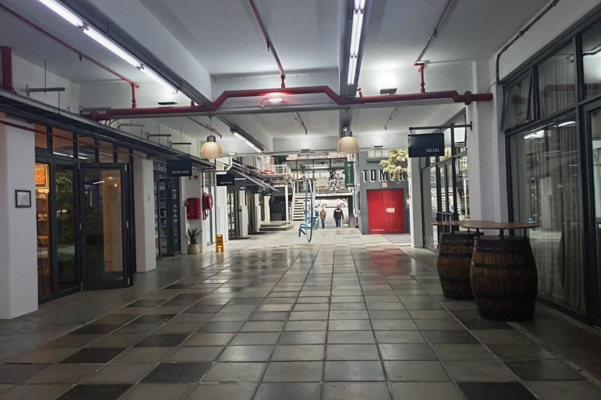 A commercial hallway or pedestrian thoroughfare featuring industrial-style exposed red fire piping, fluorescent lighting, and tiled flooring with access to various office or retail units.