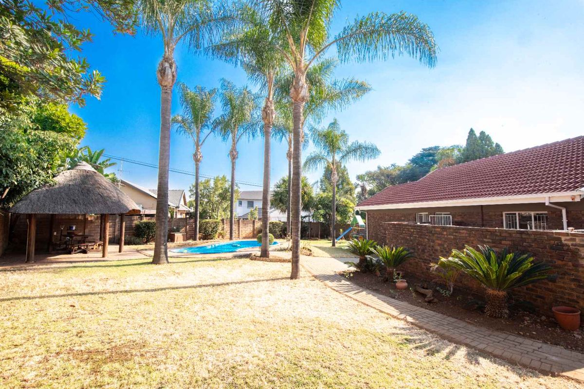 Spacious rear garden featuring a swimming pool, a traditional thatched lapa, paved pathways, and established palm trees.