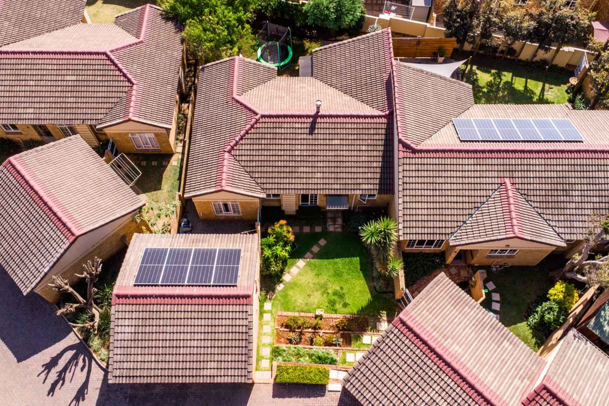 Aerial view of a cluster property featuring multiple tiled roof sections, integrated solar panels, a well-maintained garden, and paved driveway areas.