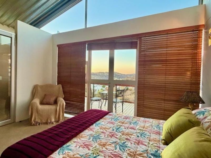 A bright bedroom featuring a sloped ceiling, large windows with timber blinds, and direct access to an en-suite bathroom and outdoor balcony.