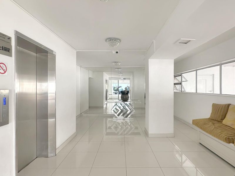 A bright and well-maintained apartment building entrance lobby featuring tiled flooring, a stainless steel elevator, and modern recessed lighting.