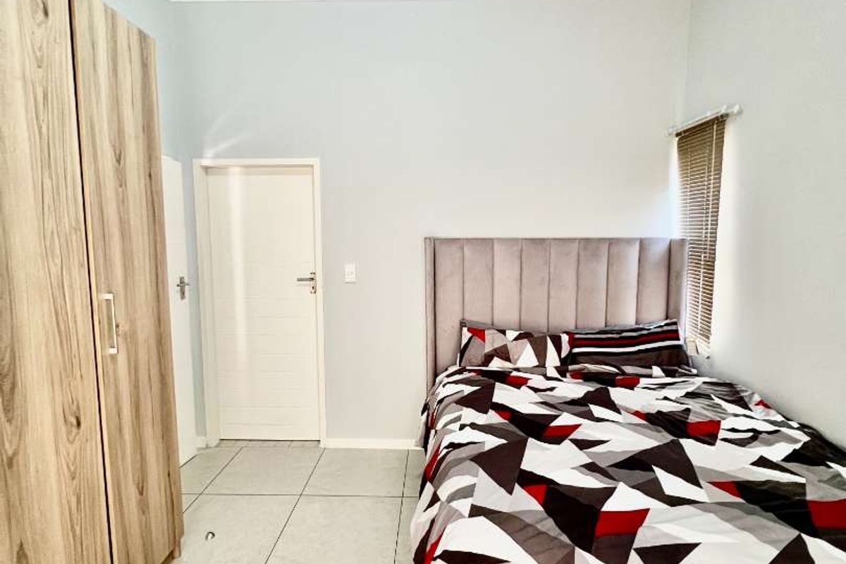 A bedroom featuring large format floor tiles, built-in wooden cupboards, and a window fitted with horizontal blinds.