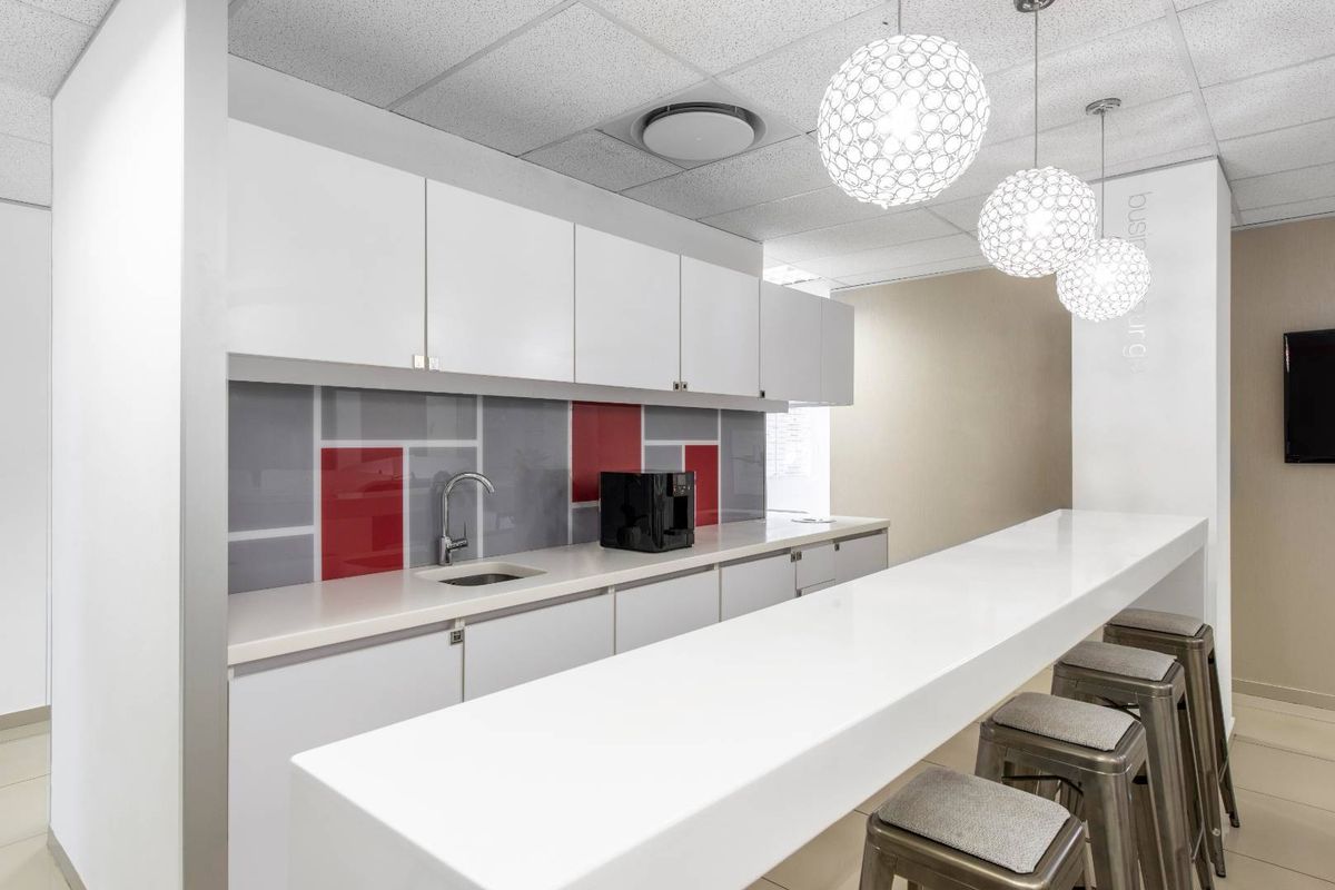 Modern office kitchenette featuring white cabinetry, a sleek countertop, a stainless steel sink, and a decorative glass splashback with red accents.