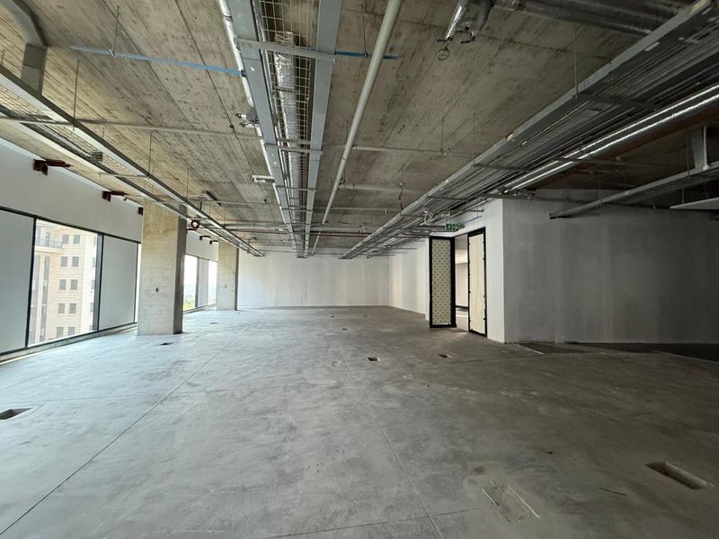 An expansive, open-plan office space in shell condition featuring exposed concrete ceilings, industrial ducting, and large windows providing ample natural light.