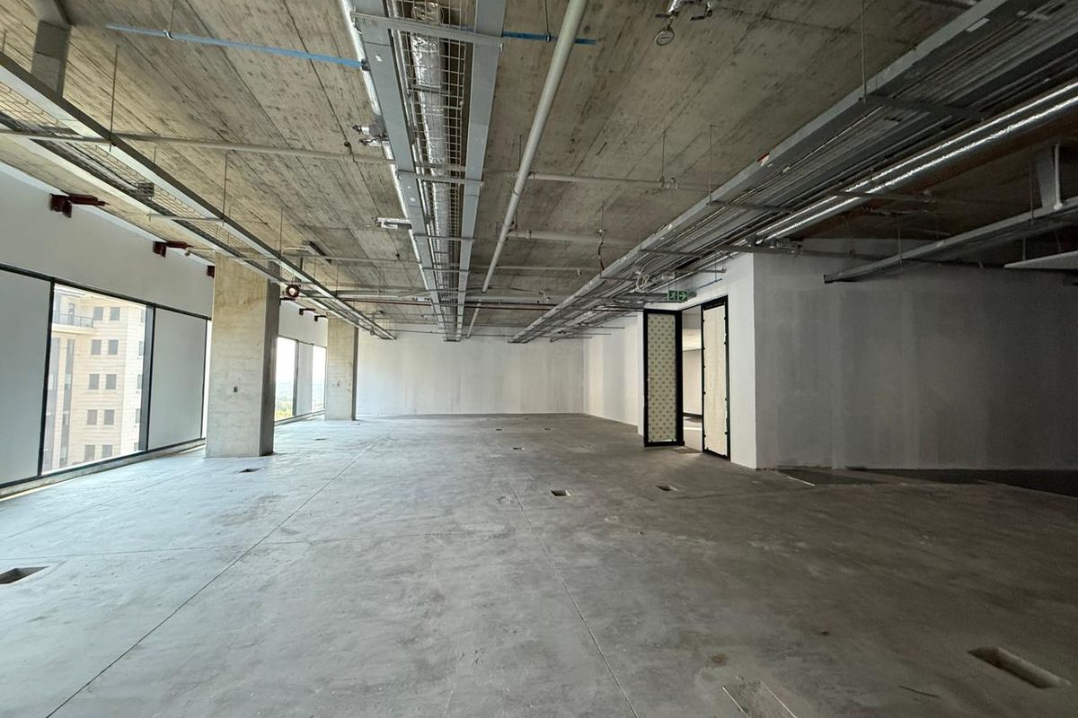 An expansive, open-plan office space in shell condition featuring exposed concrete ceilings, industrial ducting, and large windows providing ample natural light.