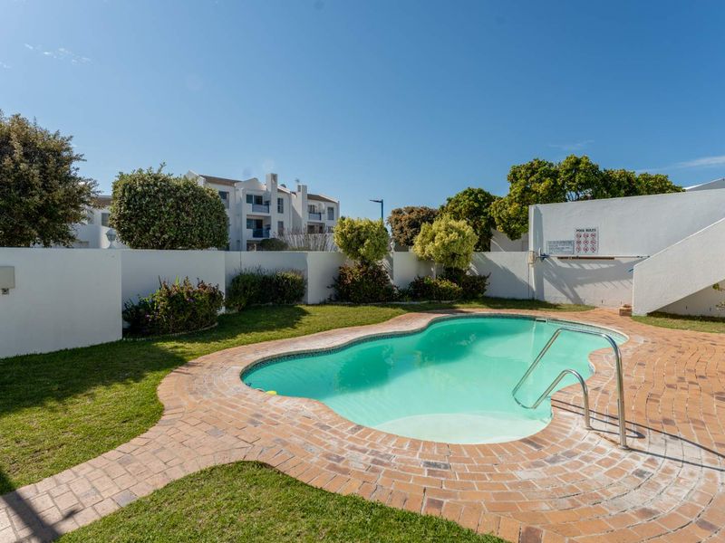 A well-maintained communal swimming pool area featuring brick paving, surrounding boundary walls, and established garden landscaping within the apartment complex.