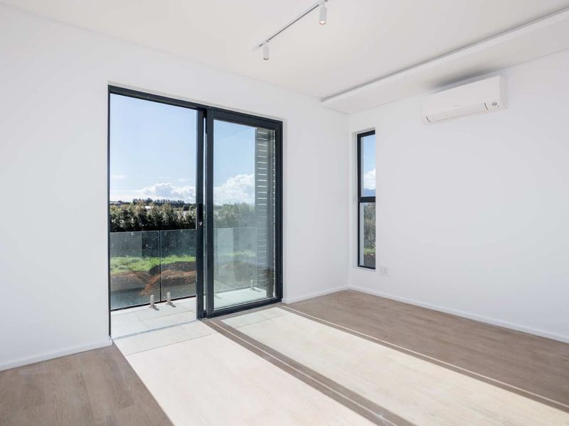 A bright, modern, empty bedroom featuring laminate flooring, track lighting, a split system air conditioner, and a sliding glass door leading to a balcony.