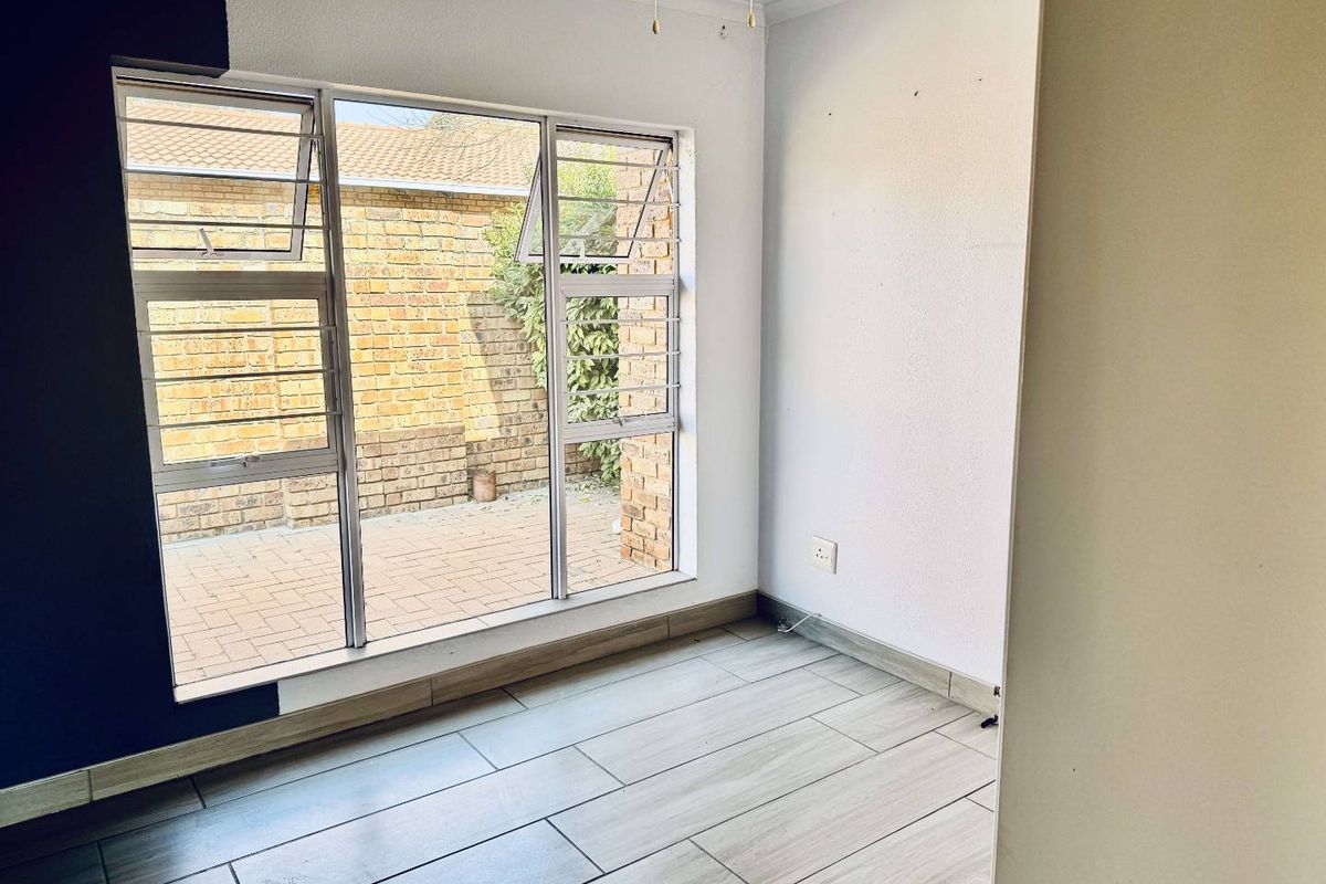 A bright, empty bedroom featuring modern wood-look floor tiles, large aluminium-framed windows, and a ceiling fan, overlooking a private paved courtyard.