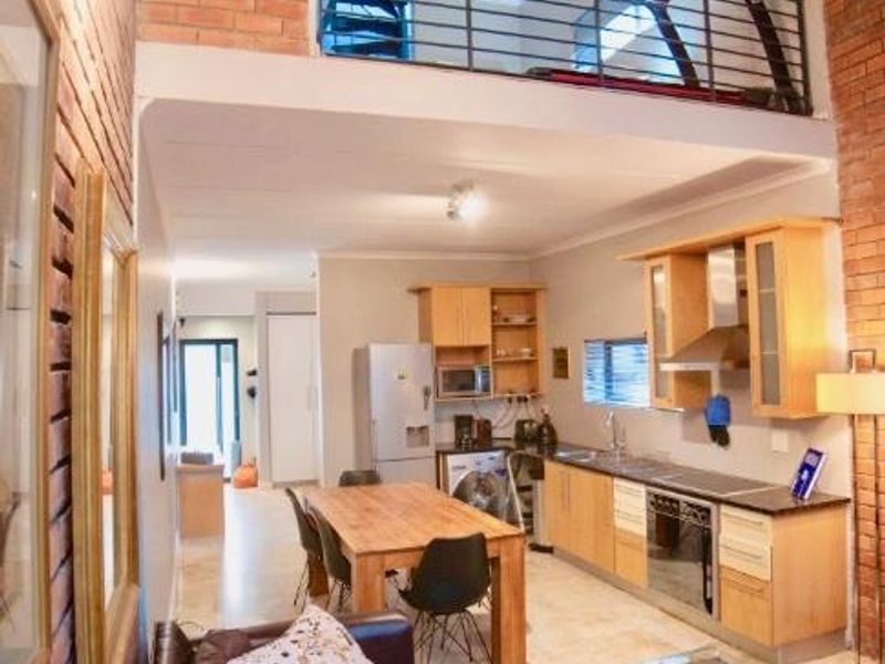 Open-plan living and kitchen area featuring exposed brick walls, a mezzanine level with metal balustrade, and functional wooden cabinetry with integrated appliances.