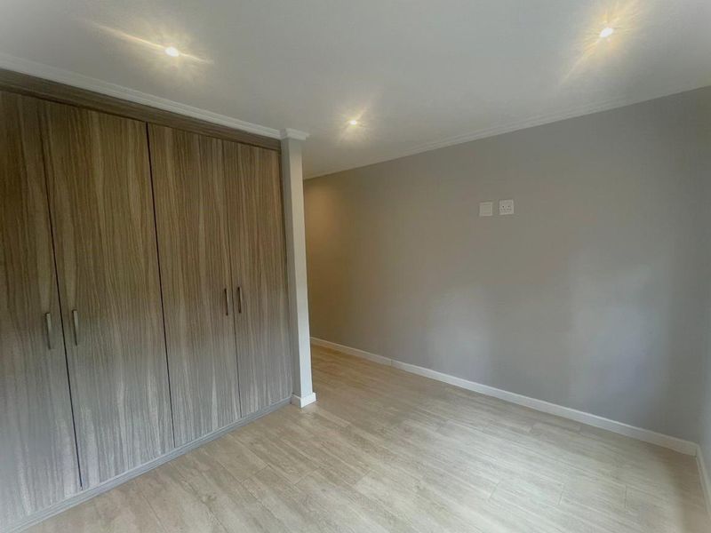 A clean, modern bedroom featuring light wood-look laminate flooring, neutral grey walls, and ample built-in cupboard storage with recessed ceiling lighting.