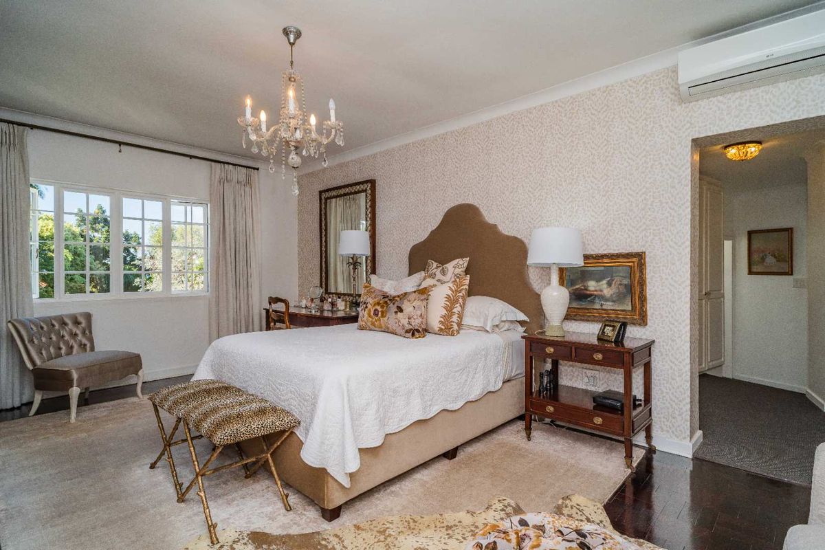 A well-appointed bedroom featuring patterned wallpaper, a crystal chandelier, and a wall-mounted air conditioning unit, with polished timber flooring.