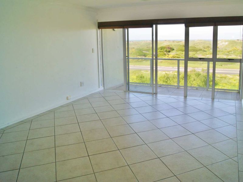 An empty, sunlit living room featuring neutral tiled flooring and large sliding glass doors that open onto a balcony with scenic coastal views.