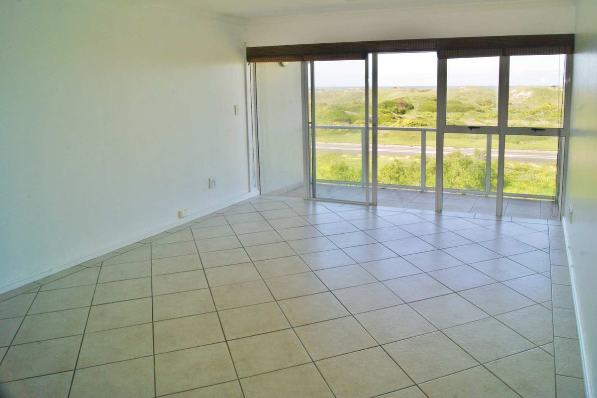 An empty, sunlit living room featuring neutral tiled flooring and large sliding glass doors that open onto a balcony with scenic coastal views.