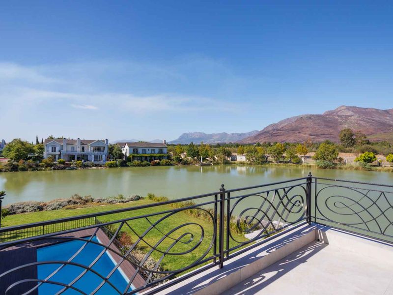 A spacious balcony featuring a decorative wrought iron balustrade, overlooking a private swimming pool, lush garden, and scenic lake with mountain views.