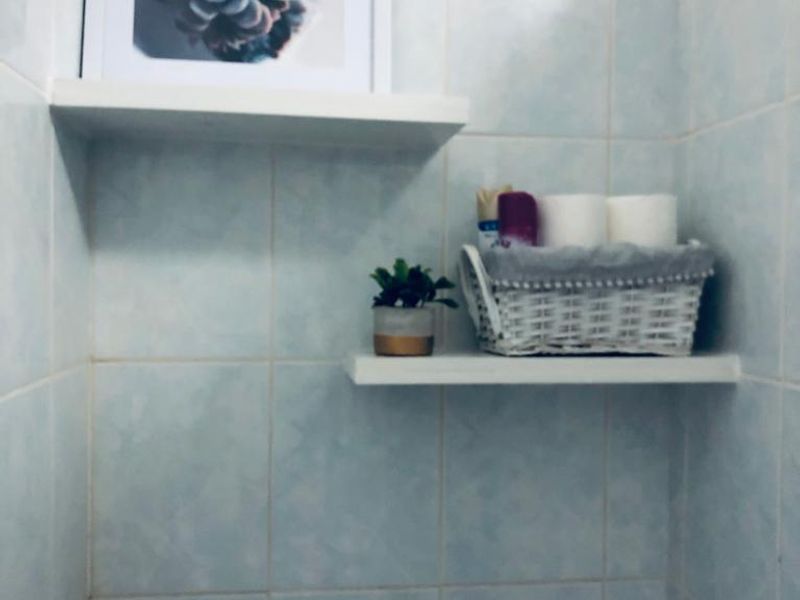 A bathroom corner featuring light blue wall tiles and two white floating shelves for storage.