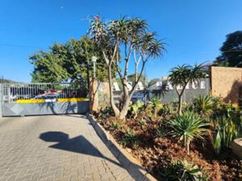 The entrance to the apartment complex features a secure gate, paved driveway, and established garden landscaping.
