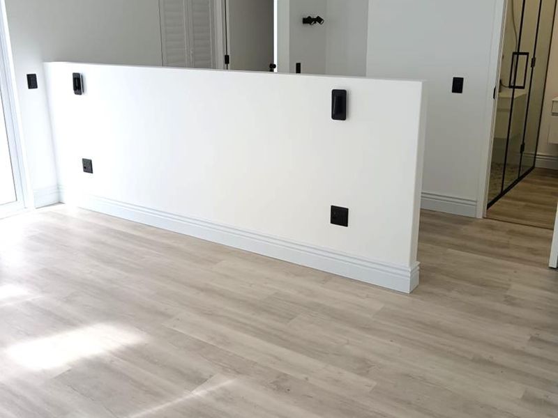 A bright, modern bedroom featuring light-toned laminate flooring, a central partition wall, and clean white finishes, offering a versatile and well-maintained living space.
