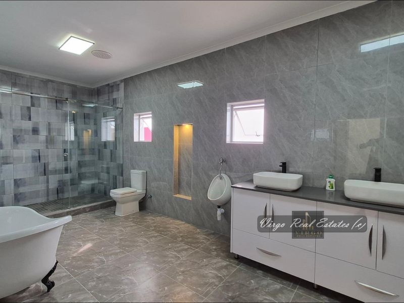 Modern, spacious bathroom featuring a freestanding bathtub, glass-enclosed shower, double vanity with vessel sinks, wall-mounted toilet, and a urinal with contemporary grey tiling.
