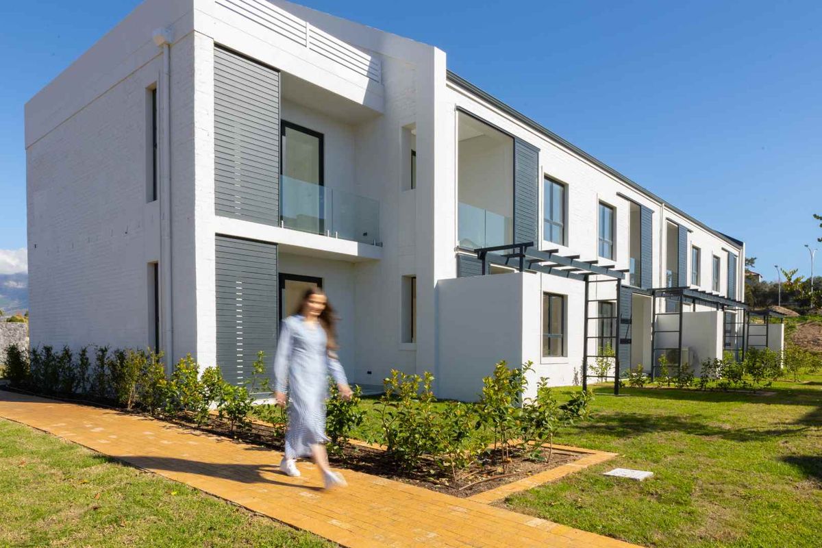 Modern apartment building exterior featuring clean white facades, grey paneling, glass balcony balustrades, and a well-maintained landscaped garden with a paved walkway.