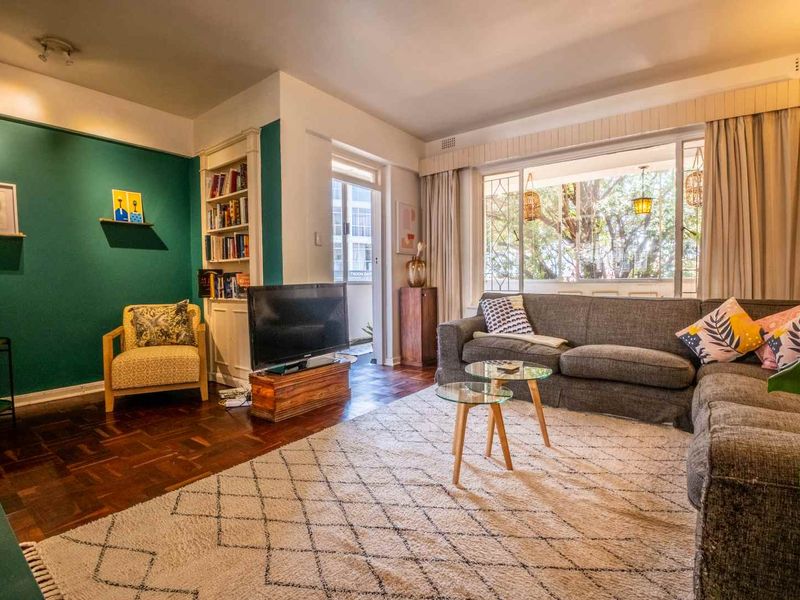 A well-lit living room featuring classic parquet flooring, a built-in bookshelf, and large windows that provide natural light and access to an outdoor area.