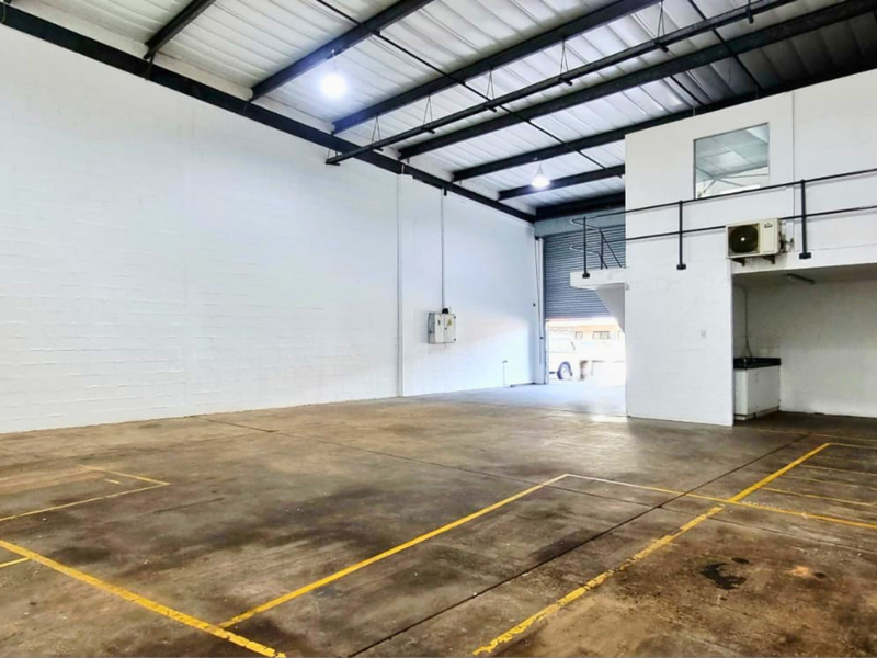 Spacious industrial warehouse unit featuring high ceilings with exposed steel trusses, a mezzanine level, concrete flooring, and a large roller shutter door for access.