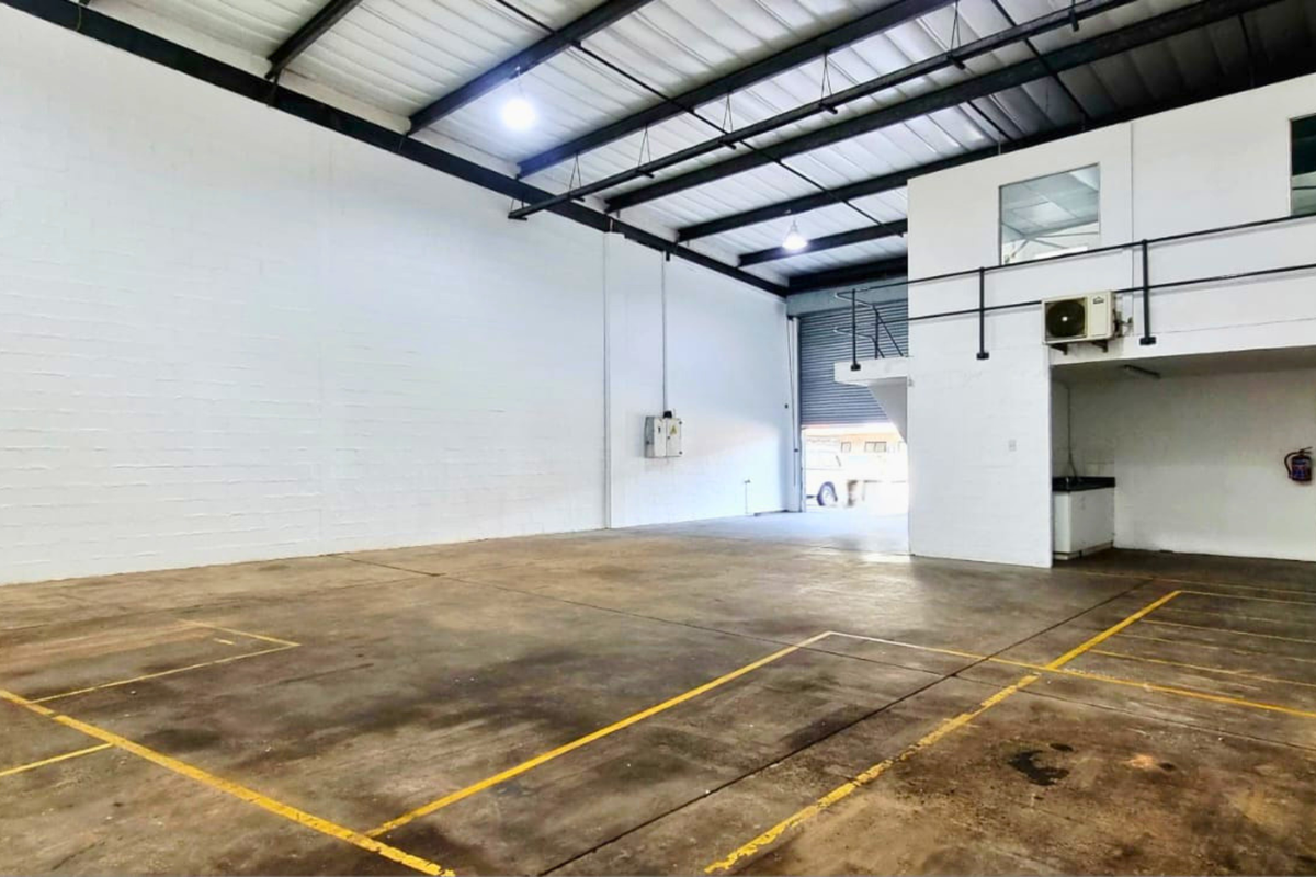 Spacious industrial warehouse unit featuring high ceilings with exposed steel trusses, a mezzanine level, concrete flooring, and a large roller shutter door for access.