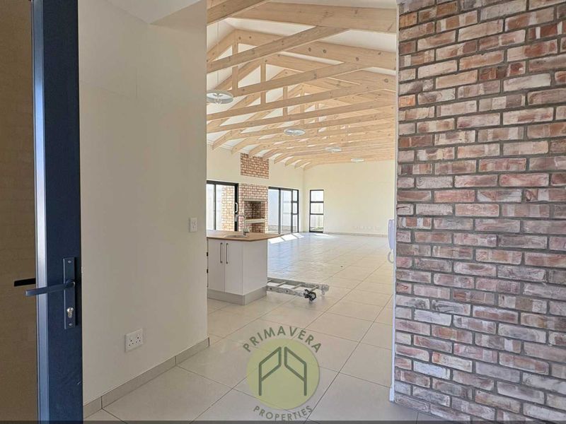 Spacious open-plan living area featuring exposed timber roof trusses, a striking brick feature wall, tiled floors, and a modern kitchen with built-in braai.