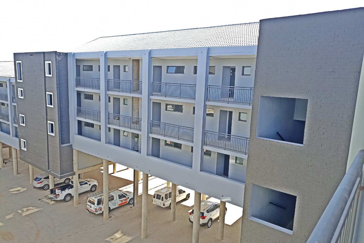 An elevated view of a modern apartment complex featuring multi-level walkways, metal railings, and covered ground-level parking supported by concrete pillars.