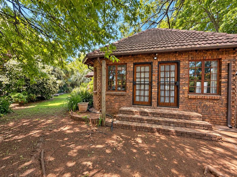A charming face brick exterior featuring a tiled roof, timber-framed windows, and brick steps leading into a lush, tree-shaded garden setting.