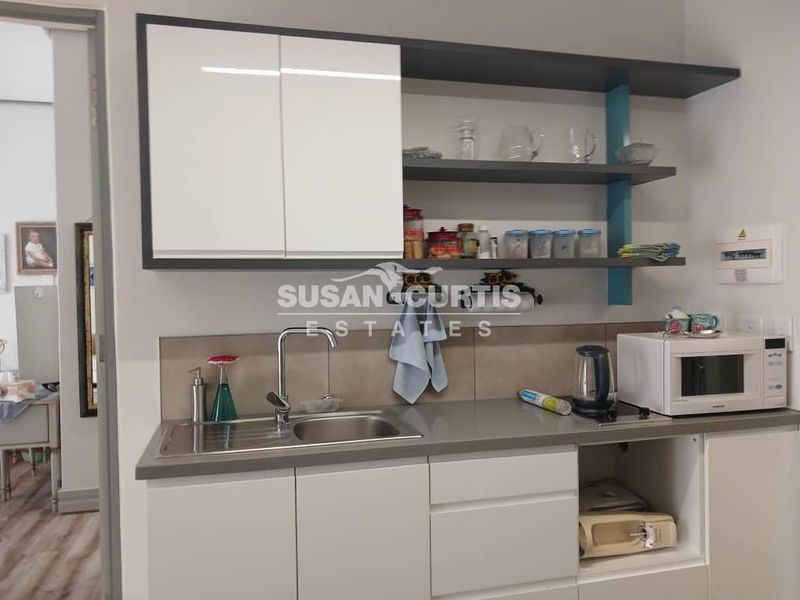 A compact, modern kitchen area featuring white cabinetry, open shelving, a stainless steel sink, and a neutral tiled splashback.