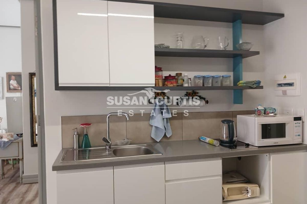 A compact, modern kitchen area featuring white cabinetry, open shelving, a stainless steel sink, and a neutral tiled splashback.
