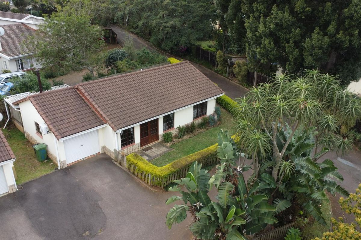 Aerial view of a townhouse featuring a tiled roof, attached garage, paved driveway, and established garden surroundings within a residential complex.