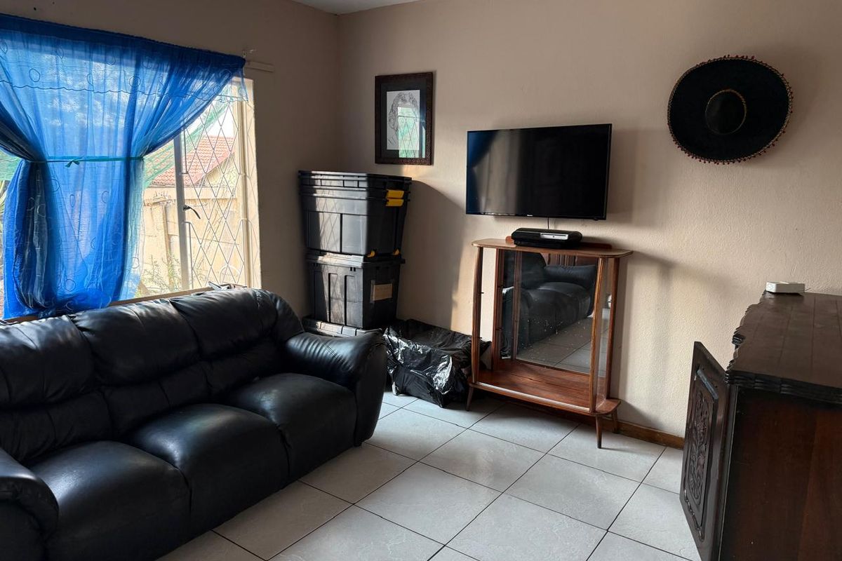 A living room featuring ceramic floor tiles, security burglar bars on the window, and a wall-mounted television.