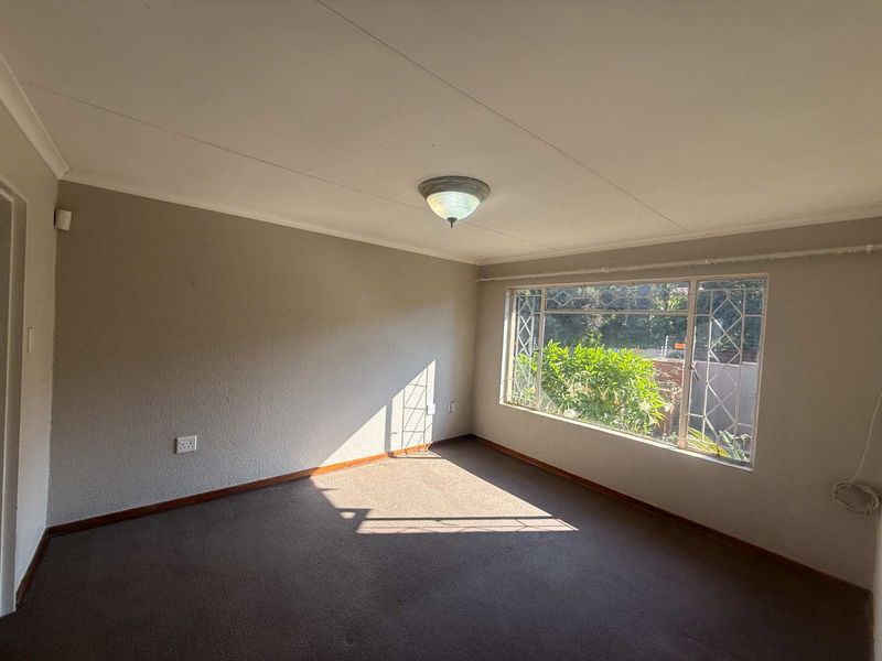 An empty living room featuring carpet flooring, a large window with security bars, and a central ceiling light fixture.