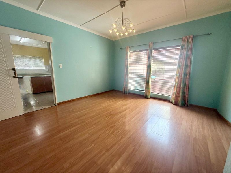 A spacious, empty living room featuring laminate flooring, a ceiling light fixture, and large windows with venetian blinds, leading into an adjacent kitchen area.