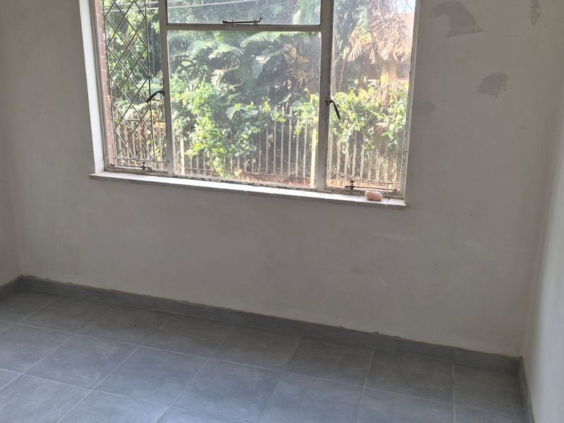 An empty bedroom featuring tiled flooring and a metal-framed window with security bars, currently requiring wall finishing and minor cosmetic updates.