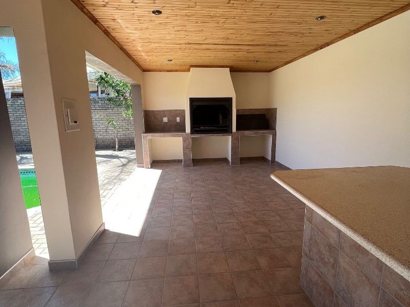 Covered outdoor patio featuring a built-in braai, stone-topped breakfast bar, tiled flooring, and a timber ceiling, overlooking the pool area.