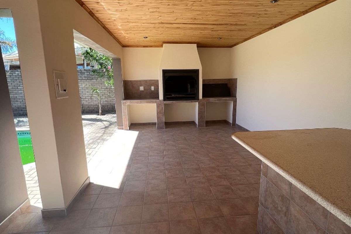 Covered outdoor patio featuring a built-in braai, stone-topped breakfast bar, tiled flooring, and a timber ceiling, overlooking the pool area.