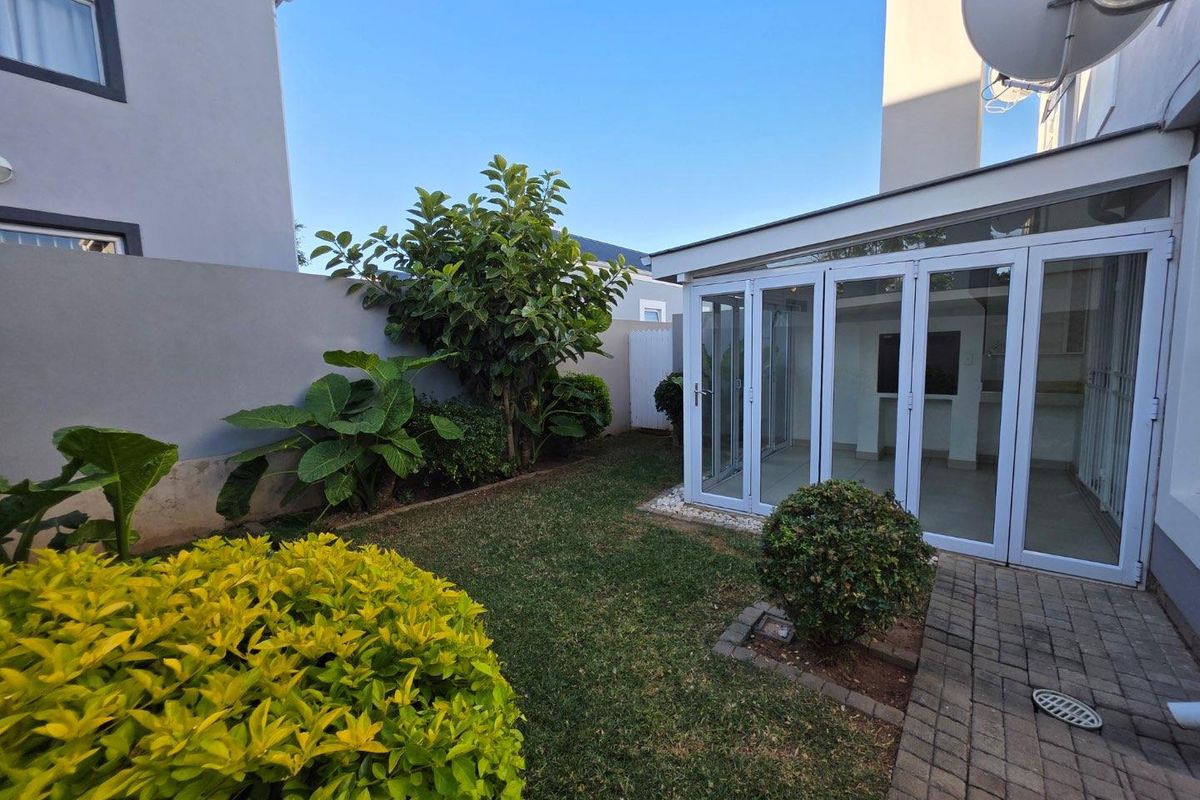 A private rear garden featuring a small lawn, landscaped shrubs, paved walkway, and a modern enclosed patio with glass folding doors.
