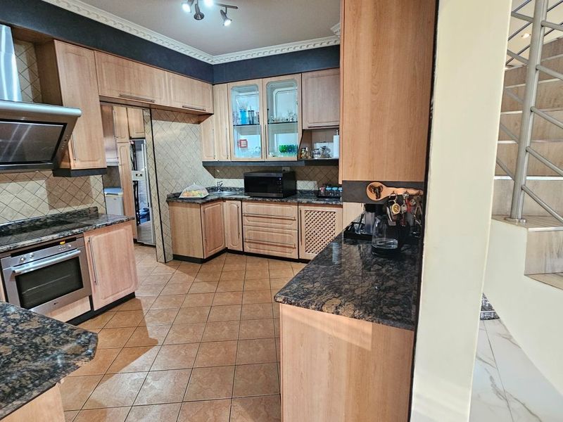 A functional kitchen featuring light wood cabinetry, granite countertops, stainless steel appliances, and tiled flooring, adjacent to an internal staircase.