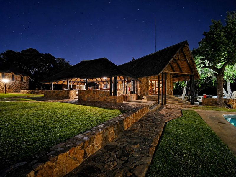 A beautifully illuminated farm-style residence at night, featuring traditional thatched roofing, natural stone masonry, and a well-maintained garden with a swimming pool.