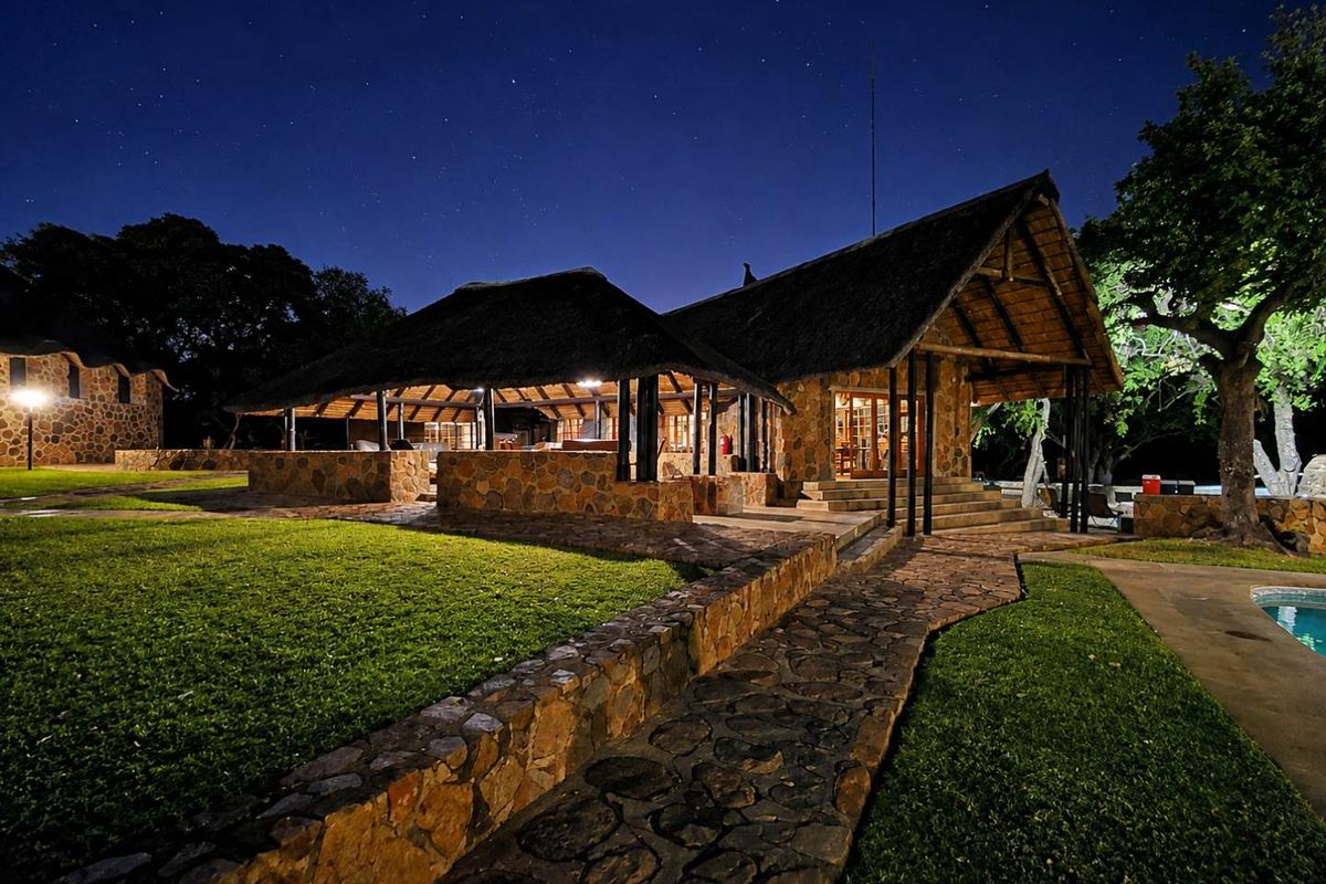 A beautifully illuminated farm-style residence at night, featuring traditional thatched roofing, natural stone masonry, and a well-maintained garden with a swimming pool.