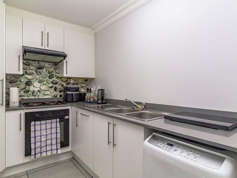 Compact kitchen featuring white cabinetry, grey countertops, a stainless steel double sink, built-in oven, extractor fan, and a decorative stone-look backsplash.