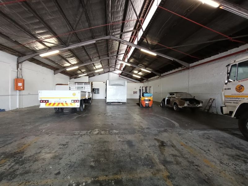 Spacious industrial warehouse featuring high ceilings with exposed steel trusses, fluorescent lighting, fire safety systems, and durable concrete flooring.