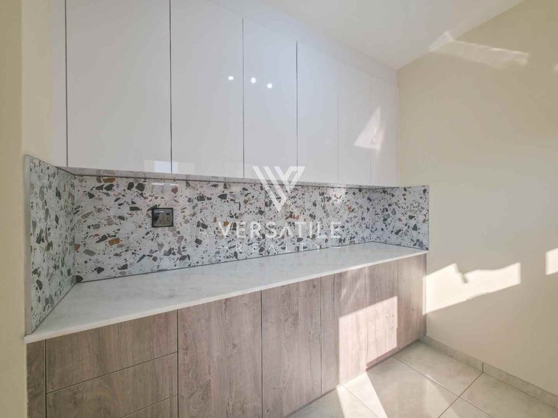 A modern, clean scullery or utility area featuring white upper cabinets, wood-grain lower cabinetry, and a stylish terrazzo-style backsplash with stone countertops.