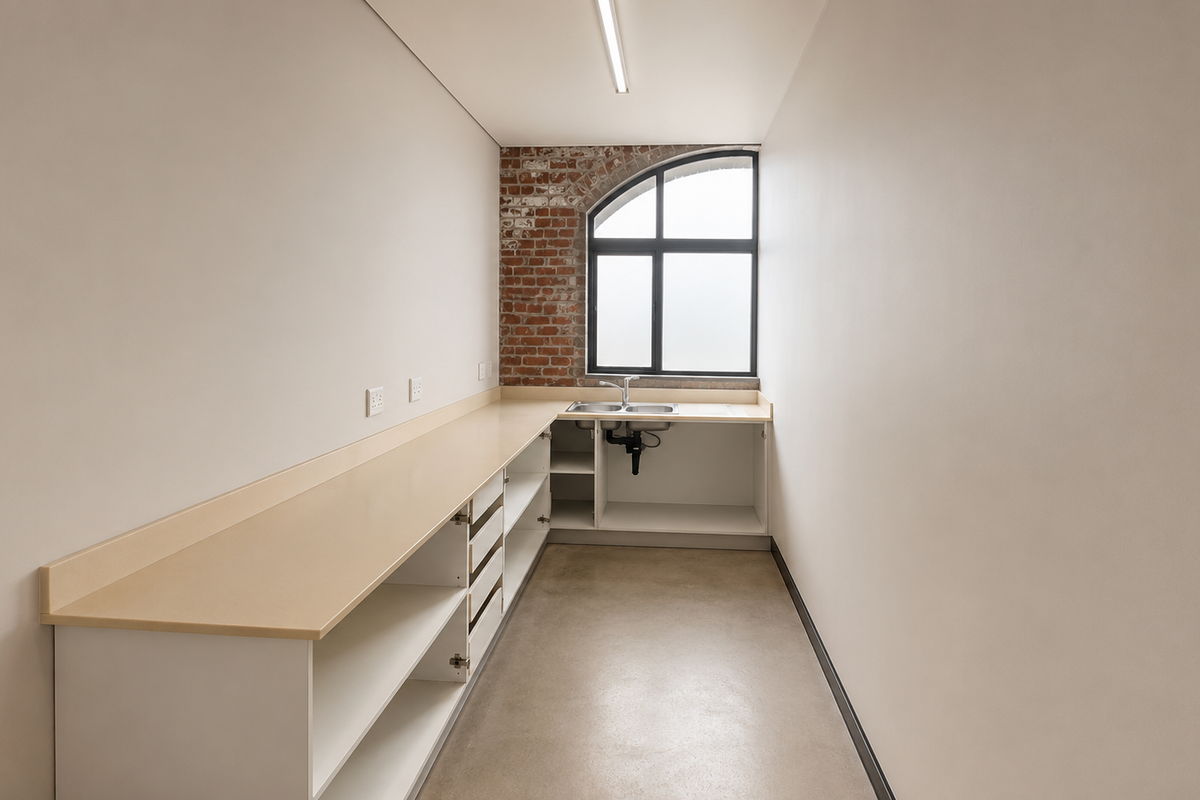 A functional office kitchenette featuring built-in cabinetry, a countertop, a stainless steel sink, and an exposed brick feature wall with an arched window.